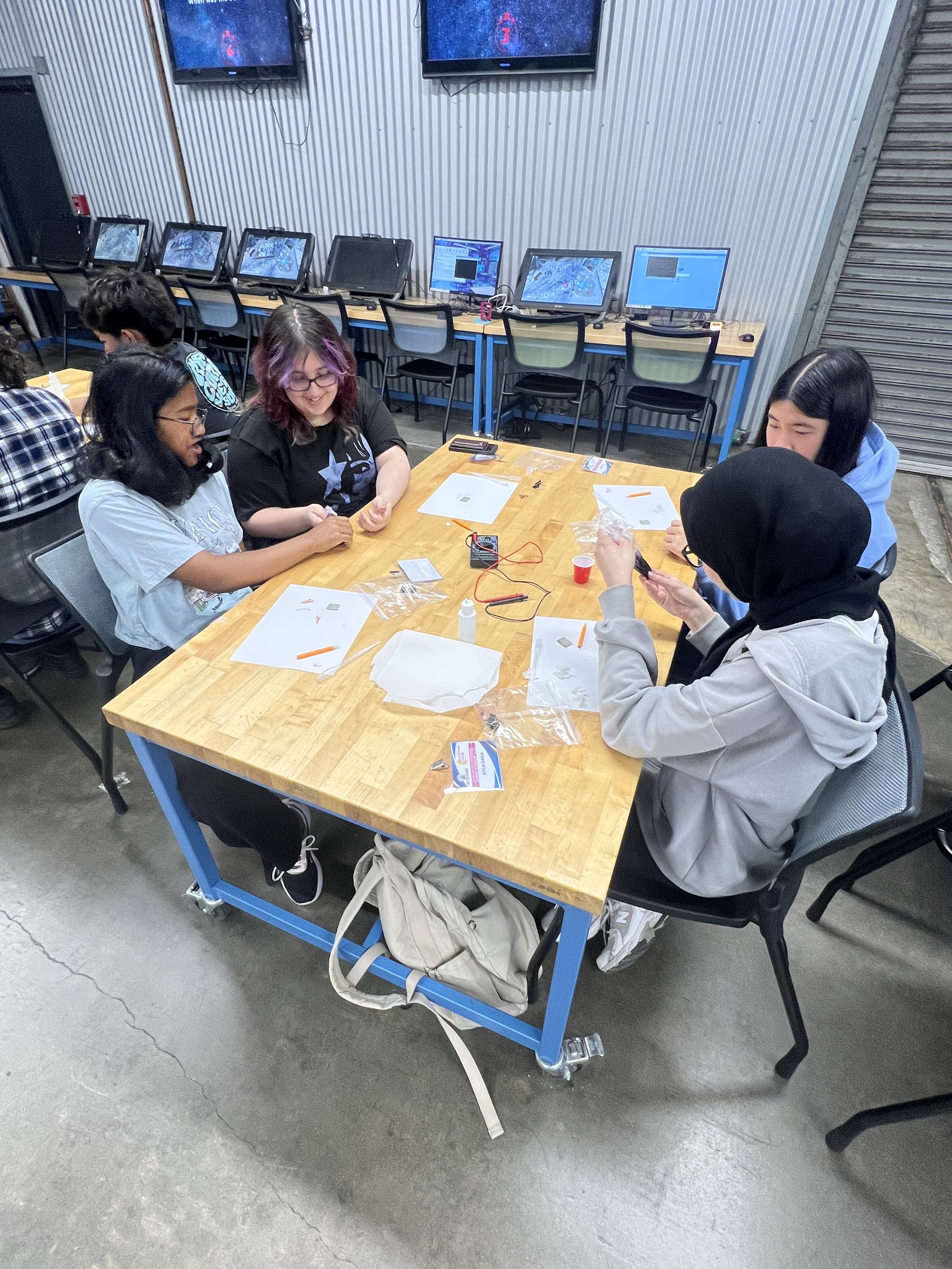 Students working at a workshop table with tools and computers in a classroom